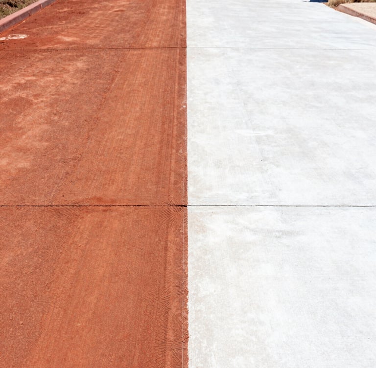 Before-and-after split view of a weathered concrete driveway in Broome, left side covered in thick red Kimberley dirt, right side blasted clean to pristine white concrete under harsh afternoon sun, 35mm