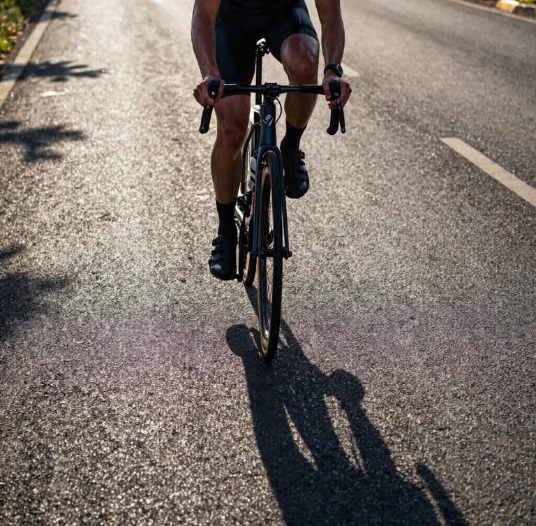 Low-angle hero shot of a cyclist in intense effort, sweat on skin, early morning light casting long dramatic shadows on the asphalt