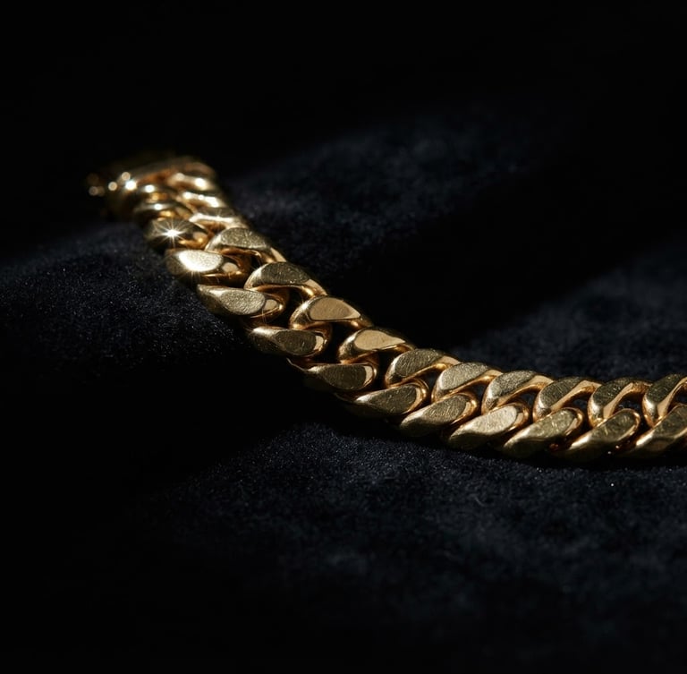 Wide panoramic shot of a heavy gold link bracelet on a matte black velvet surface, dramatic low-key studio lighting, sharp metallic gold highlights glinting from the left third, emerging from deep shadows, expansive dark background.