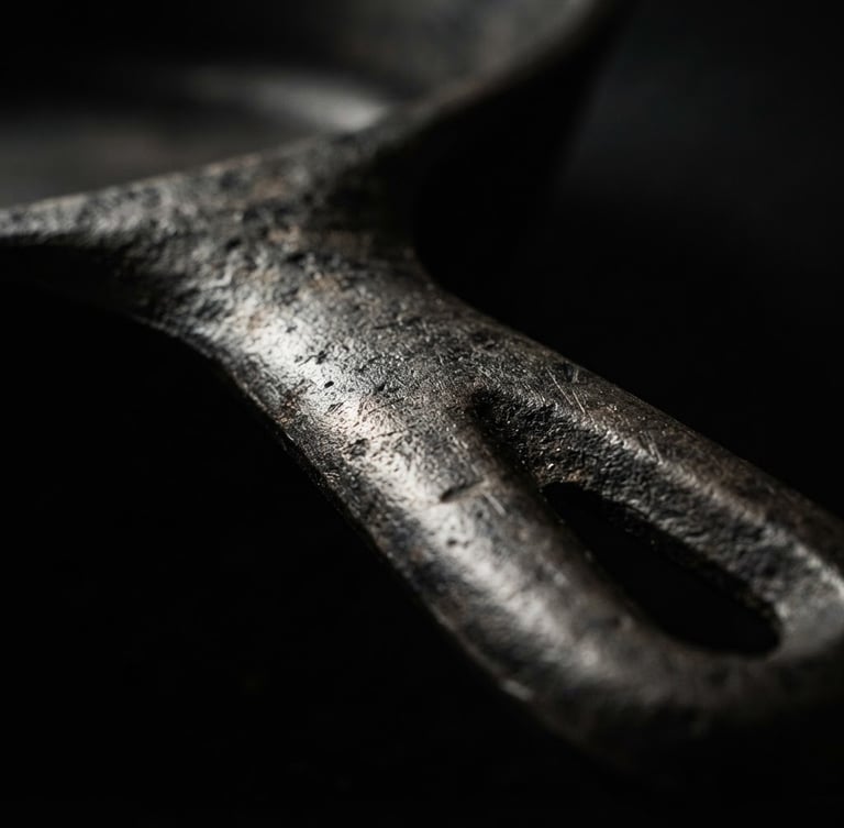 Extreme close-up macro shot of a heavy unlacquered iron skillet handle, dramatic side-lighting, deep shadows, dark moody background, high-contrast tactile metal textures