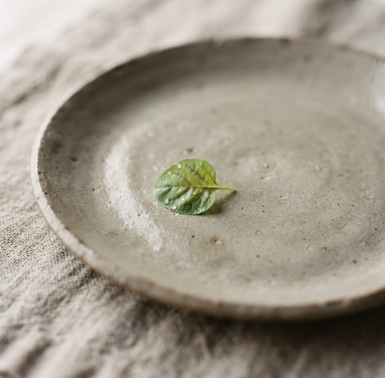 Extreme close-up of a minimalist ceramic plate with a single micro-green leaf, soft natural side light, raw linen napkin texture, muted warm gray tones, shallow depth of field, 35mm film aesthetic