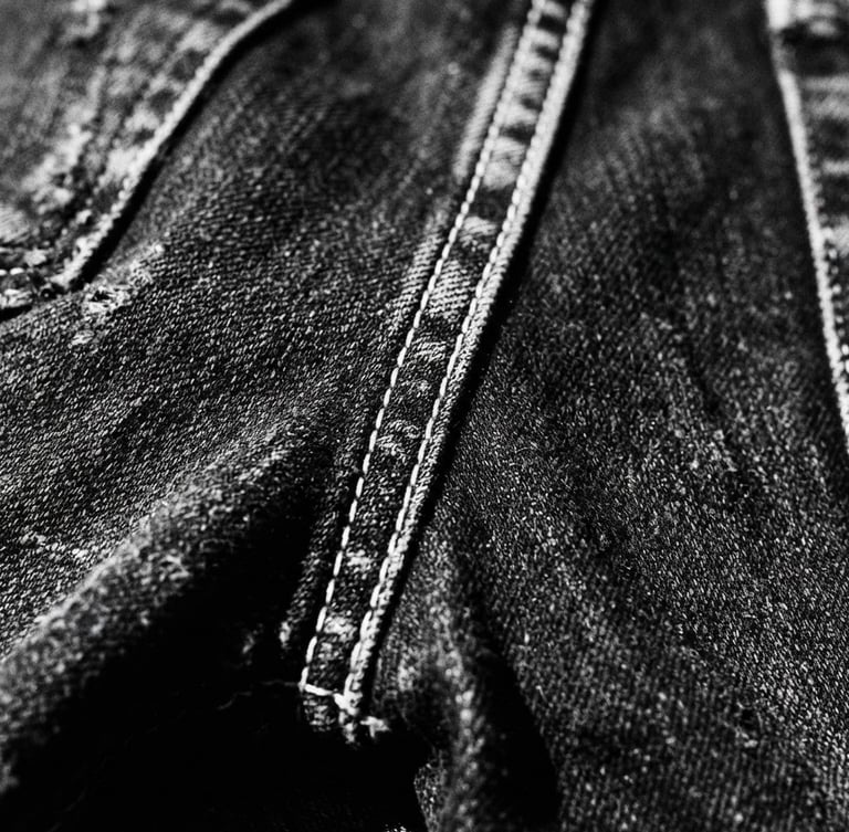 Close-up of distressed black denim fabric with visible white stitching, harsh direct sunlight, deep shadows, high-contrast monochrome, 35mm film grain, side view, wide angle.