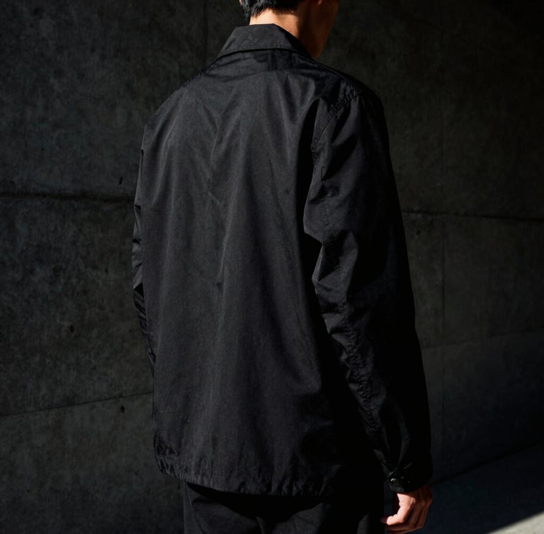 Low-key, high-contrast model shot from behind, showing the architectural drape of a structural black jacket against a dark concrete wall, harsh side-lighting, 35mm film