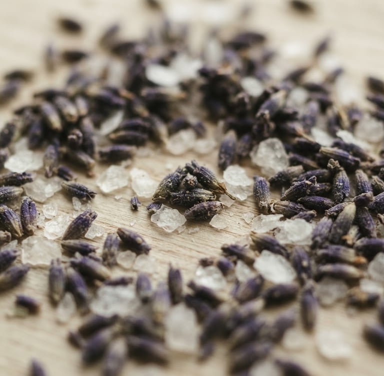 Macro close-up of dried lavender buds and magnesium flakes on a light ash wood surface, soft diffused daylight, shallow depth of field, muted sage and cream tones.