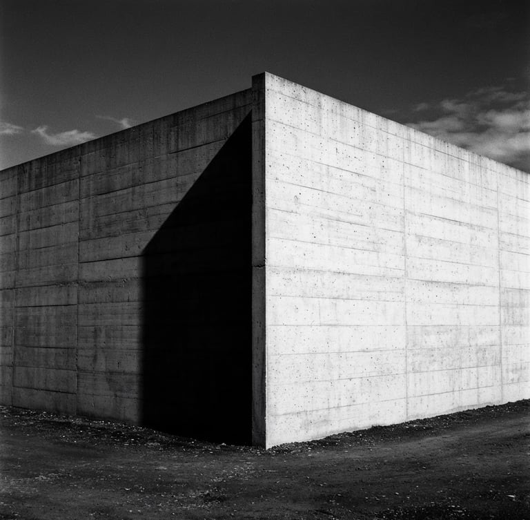 Minimalist architectural photography. Stark concrete corner of a Swiss modernist building under harsh direct sunlight, casting a sharp, deep geometric shadow. High-contrast monochrome, 35mm lens.
