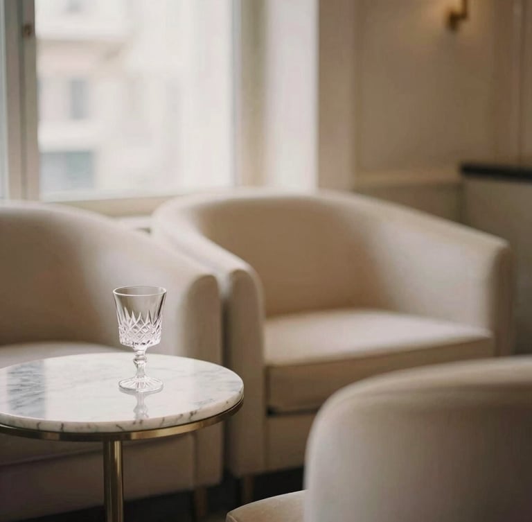 Editorial portrait of a sophisticated private VIP lounge with minimalist champagne-colored furniture, a marble side table with a crystal glass, soft warm window light, elegant atmosphere, shallow depth of field.