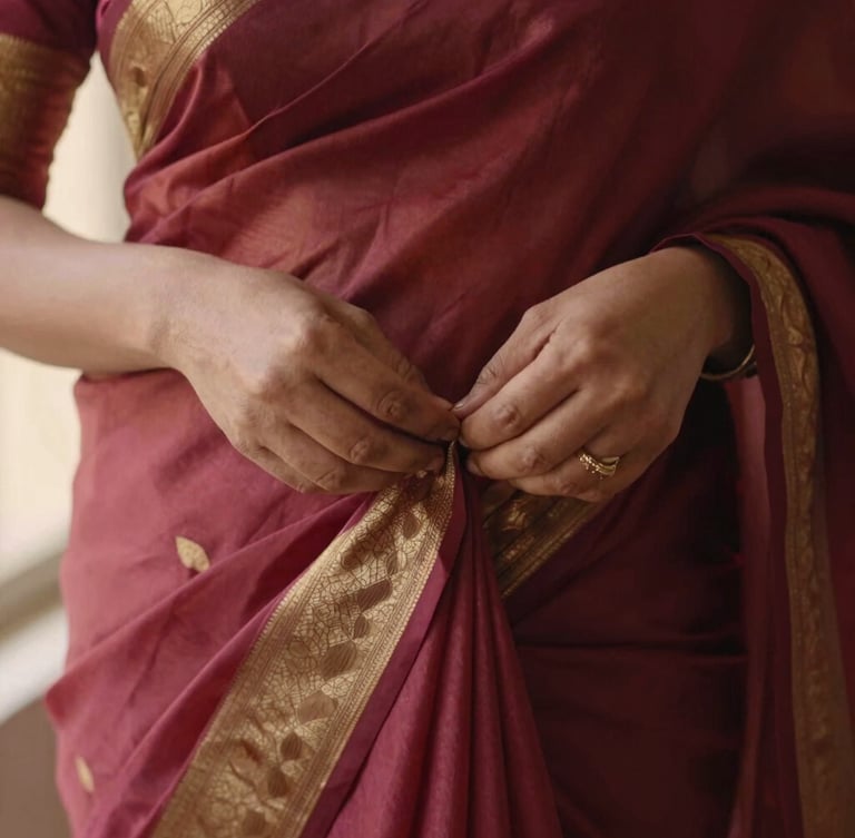 Close-up of hands meticulously pleating a rich crimson and gold silk saree, warm diffused lighting, editorial aesthetic, 35mm