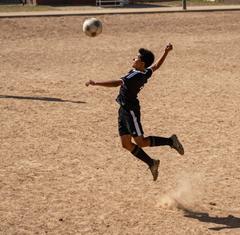 Low-angle heroic shot of a young player mid-air striking the ball on a dirt pitch, harsh midday sun casting deep shadows, raw high-stakes athletic intensity, gritty sports photography.