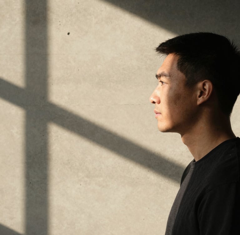 Portrait of a creative professional in profile, harsh direct sunlight casting a sharp shadow of window frames onto a warm concrete wall, minimalist composition, warm tones, subtle grain.
