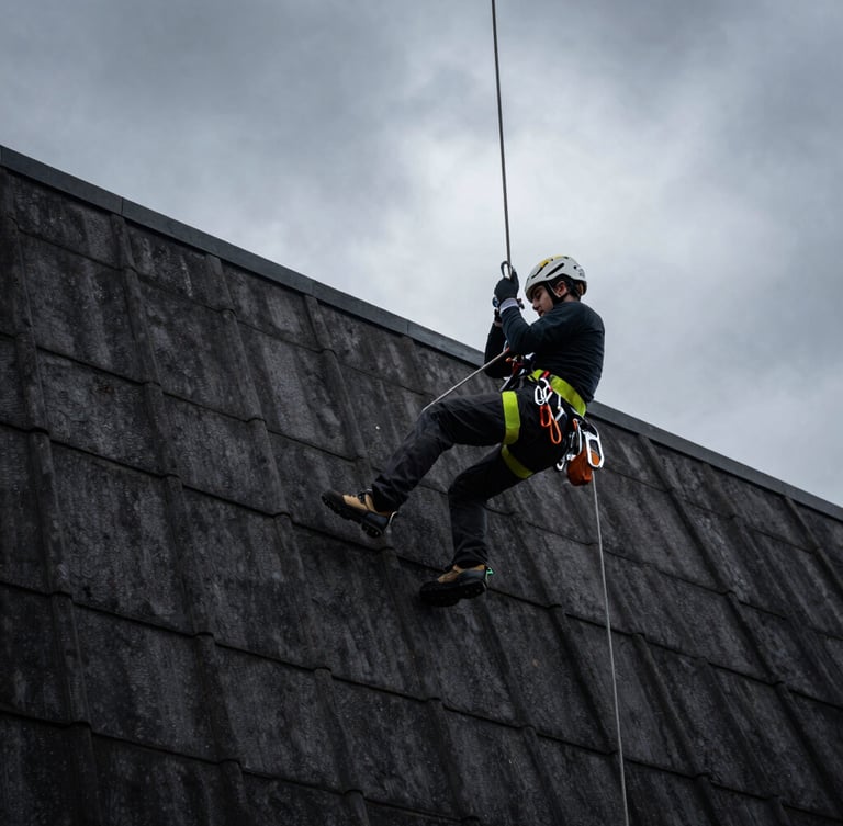 Low-angle dynamic shot of a professional industrial climber in high-visibility safety harness suspended on ropes against a dark tiled roof, dramatic overcast sky, sharp focus on technical carabiners.