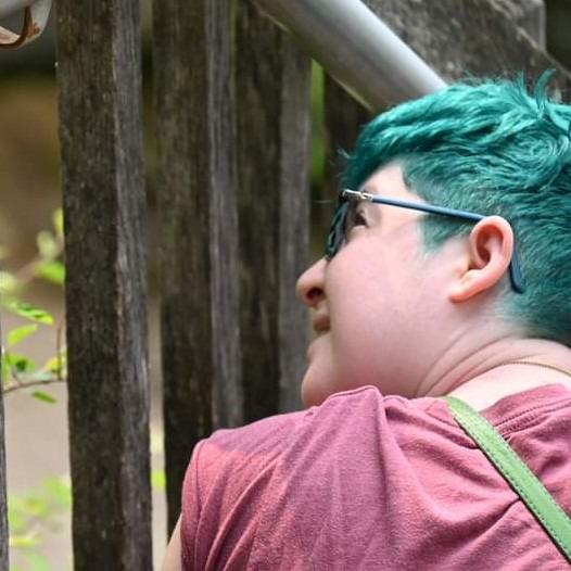 An image of the artist from behind, looking up and to the left. They have short teal hair.