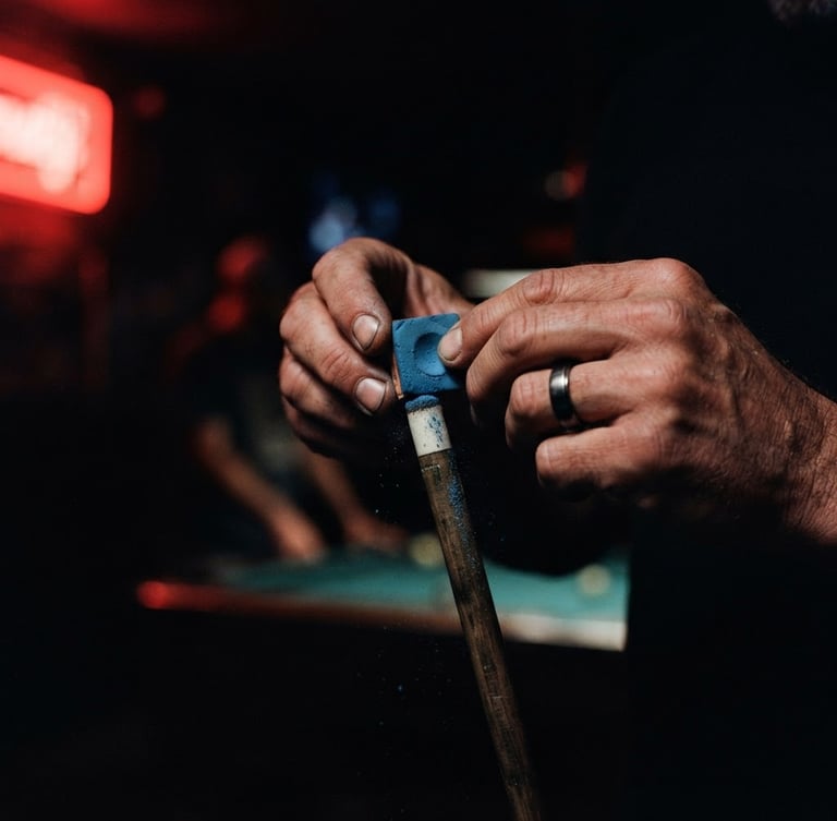 Moody low-key close-up of hands chalking a pool cue under low red neon light, dark background, cinematic 35mm photography