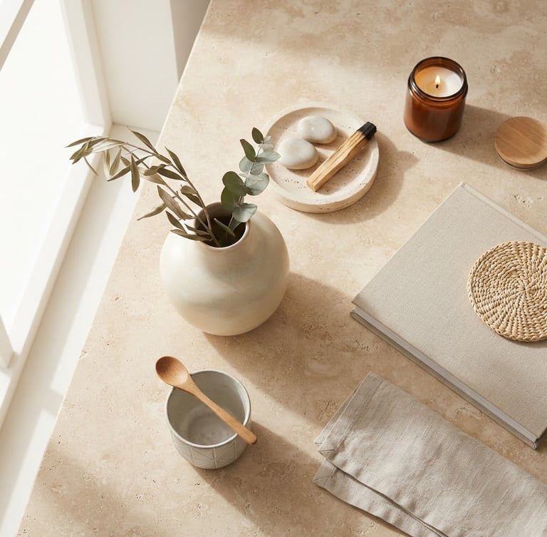Overhead styled flat-lay of minimalist home decor accessories on a warm stone surface, bright natural window light, soft diffused shadows