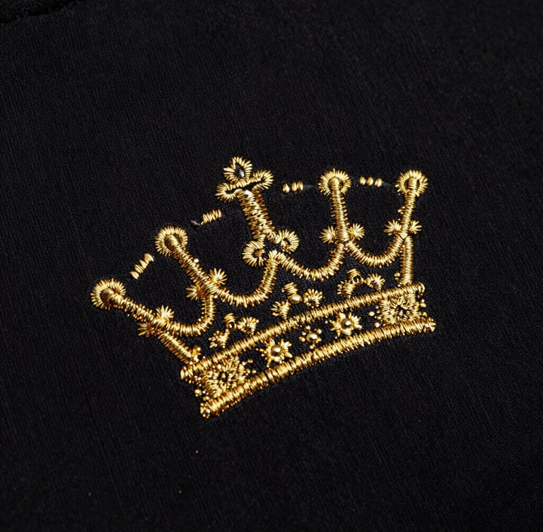 Close-up of premium heavy black cotton fabric texture, detailed gold embroidery of a crown, high-contrast dark lighting, sharp metallic reflections, 35mm