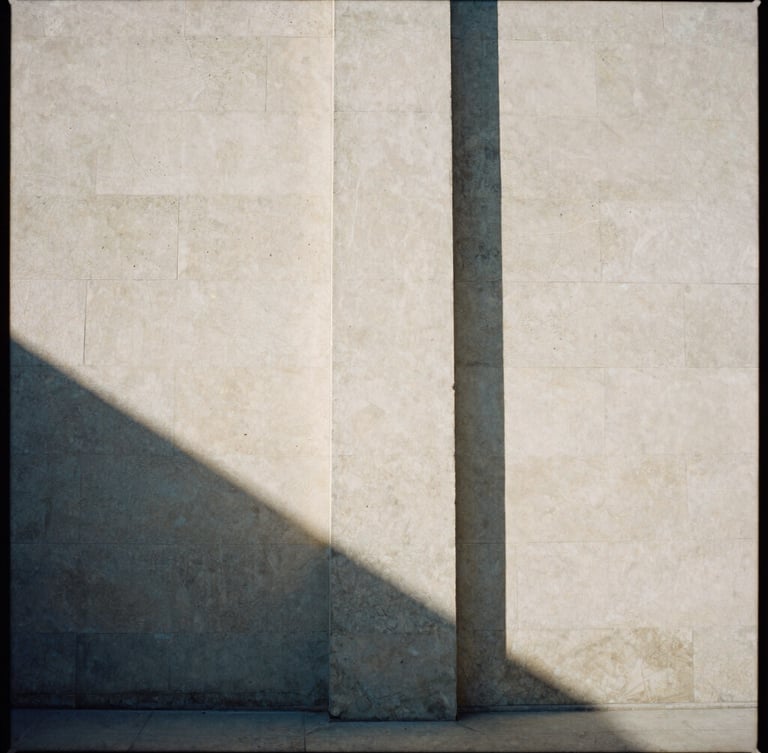 Stark architectural concrete pillar casting a long, sharp shadow on a warm stone wall, high-contrast natural light, minimalist editorial style, 35mm film grain