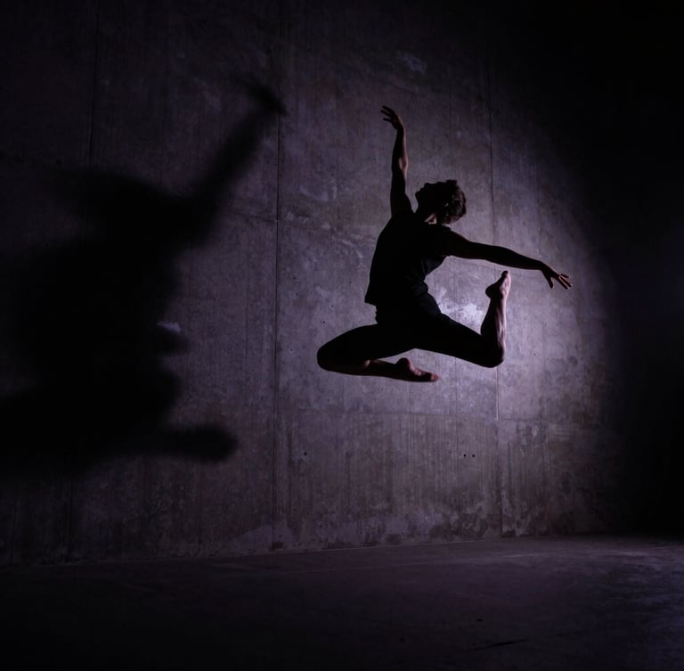 Dramatic low-angle cinematic editorial photography of a dancer in mid-air silhouette against a dark, high-contrast backlit concrete wall, deep shadows, 35mm anamorphic lens.