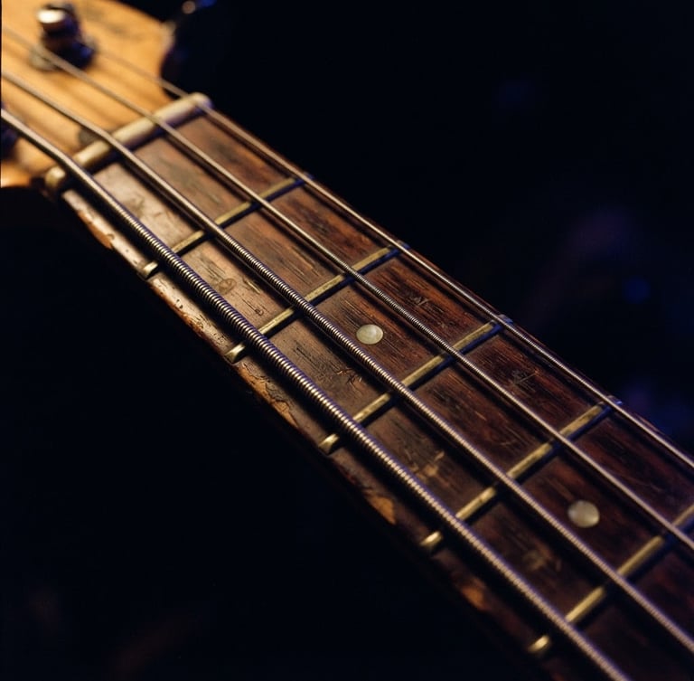 Extreme close-up of a vintage bass guitar neck, warm tungsten glow on worn wood and metal strings, deep obsidian background, 35mm film grain.