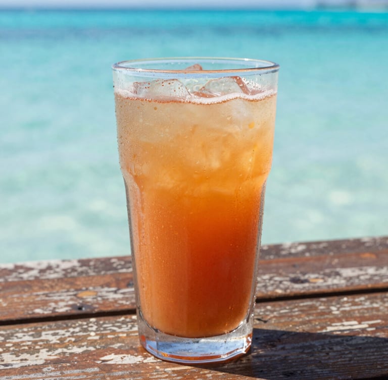 A close-up details shot of a cold tropical drink with condensation on the glass, sitting on a weathered wooden table with bright turquoise ocean water blurred in the background, warm golden sunlight