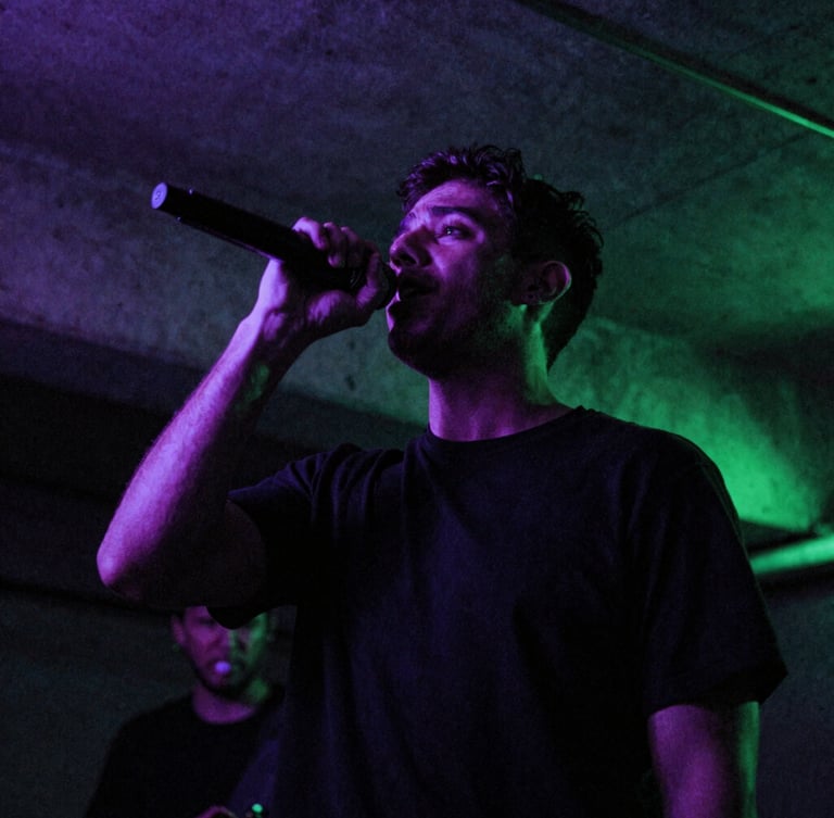 Low-angle gritty portrait of SergioMdrz performing in a dark concrete basement, glowing violet and neon green gel lights, motion blur, 35mm