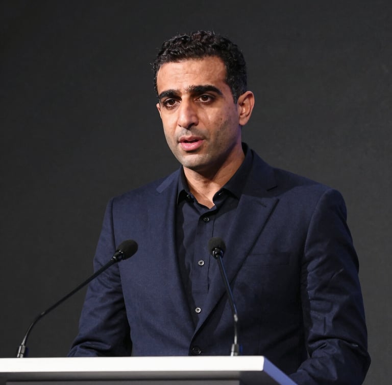 Medium-close environmental portrait of Karim speaking at a high-level conference podium, crisp natural lighting, deep navy tones, 35mm