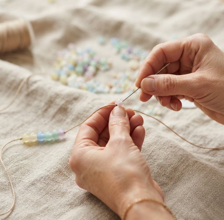 Macro close-up of hands mid-assembly, threading soft pastel glass beads onto a cord over a textured linen cloth, warm diffused daylight