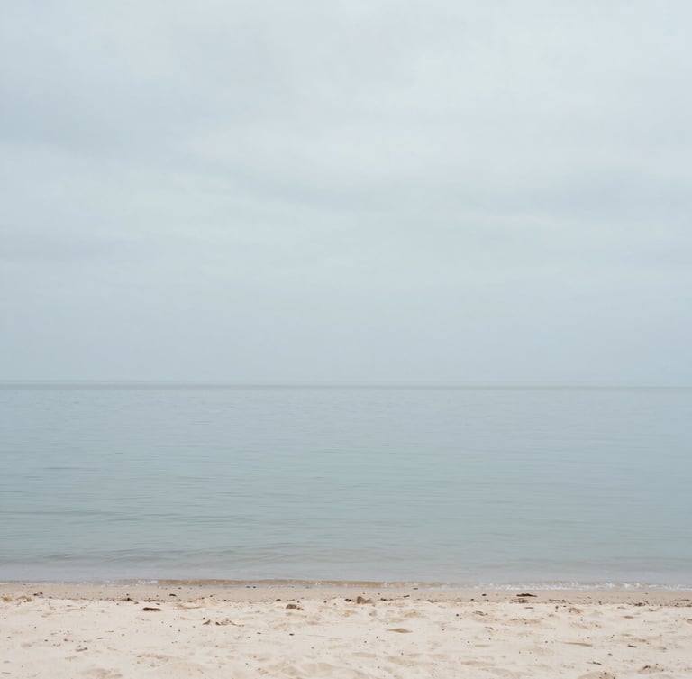 Tranquil coastal shoreline with calm water, soft overcast gray sky, pale sand, minimalist editorial photography, 35mm lens
