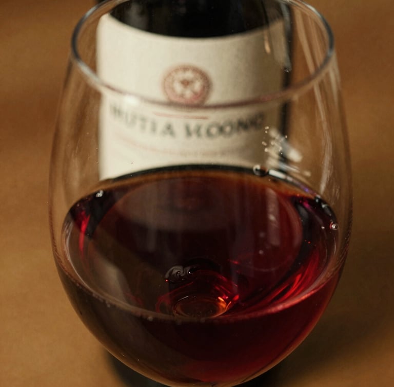 Macro shot of red wine swirling in a clear glass, warm golden hour light, rich burgundy tones, textured paper label of a wine bottle slightly visible in the background, 35mm film look.