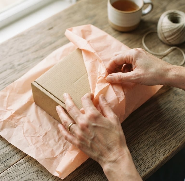 Overhead flat-lay of hands carefully wrapping a textured cardboard gift box in soft peach tissue paper, natural window light, muted organic tones, shallow depth of field