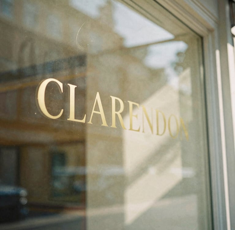 Intimate close-up of a sunlit Clarendon storefront window with gold lettering, soft morning light reflecting on glass, warm shadows, 35mm film look