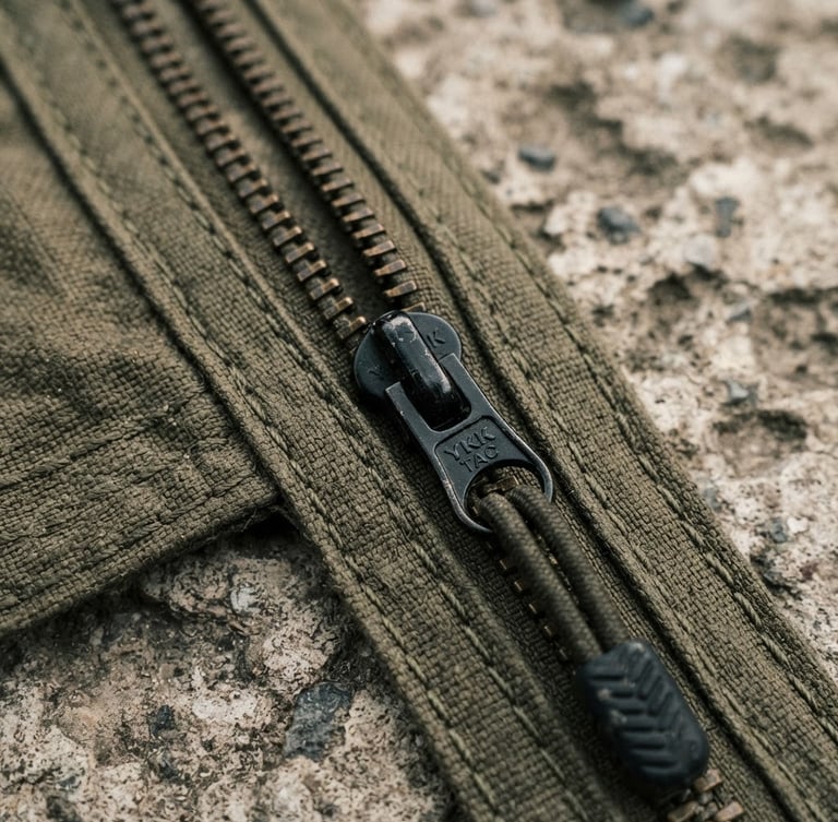 Macro close-up of heavy-duty tactical zippers and olive-drab double-stitched seams on weathered concrete, hard overcast daylight, desaturated earth tones, 35mm