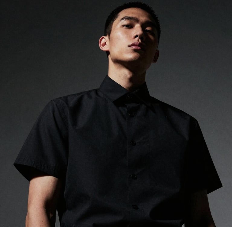 Dramatic low-angle portrait of a model wearing a structured matte black short-sleeve shirt, high-contrast directional lighting, deep shadows, desaturated tones, 35mm film grain