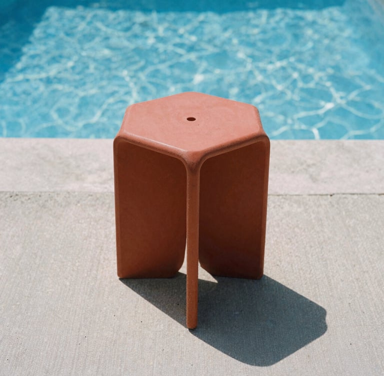 Low-angle architectural shot of a terracotta red monobloc stool casting a sharp shadow on a raw concrete pool deck under harsh midday sun, sparkling blue water in the background, crisp architectural photography.