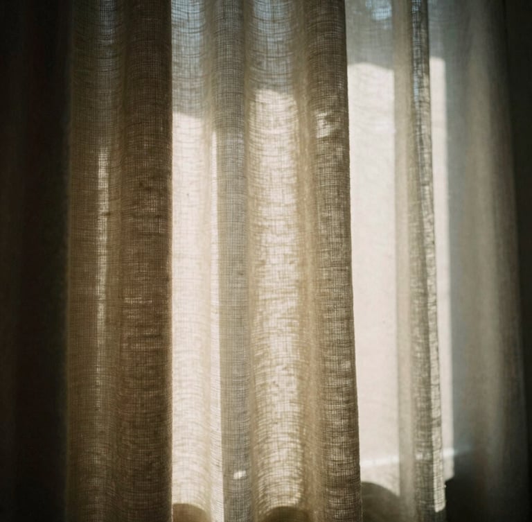 Warm editorial photograph, dramatic window light casting sharp diagonal shadows across a textured linen blackout curtain, shallow depth of field, 35mm