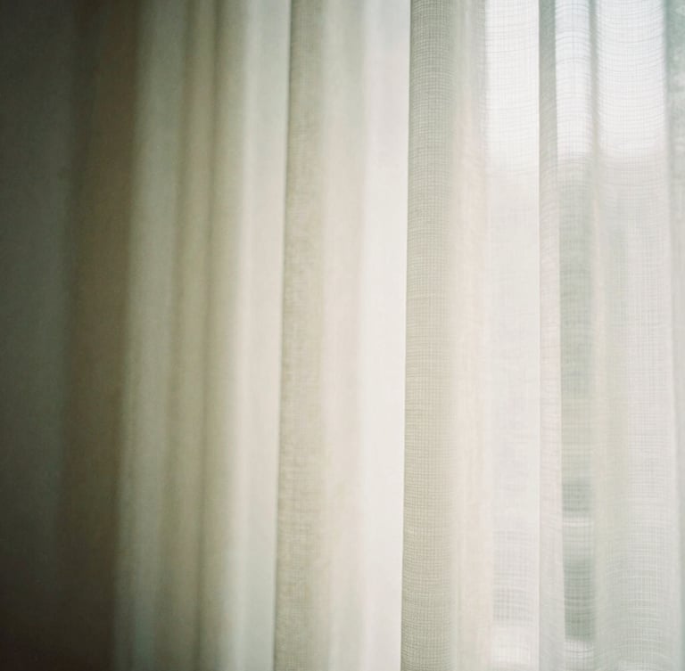 Soft architectural detail showing gentle morning light entering a quiet room through a sheer curtain, warm cream and sage green tones, shallow depth of field, 35mm