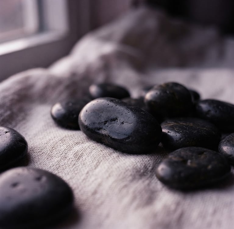Macro shot of smooth dark basalt stones resting on textured clean linen, soft twilight window light with gentle lilac undertones, shallow depth of field.