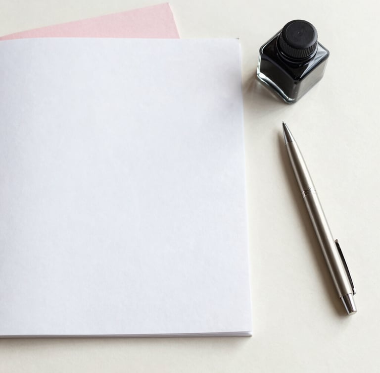 Overhead flat-lay of heavy-weight cotton paper stock, a modern glass ink well, and a single metal pen resting on a clean cream plaster surface under soft morning light.