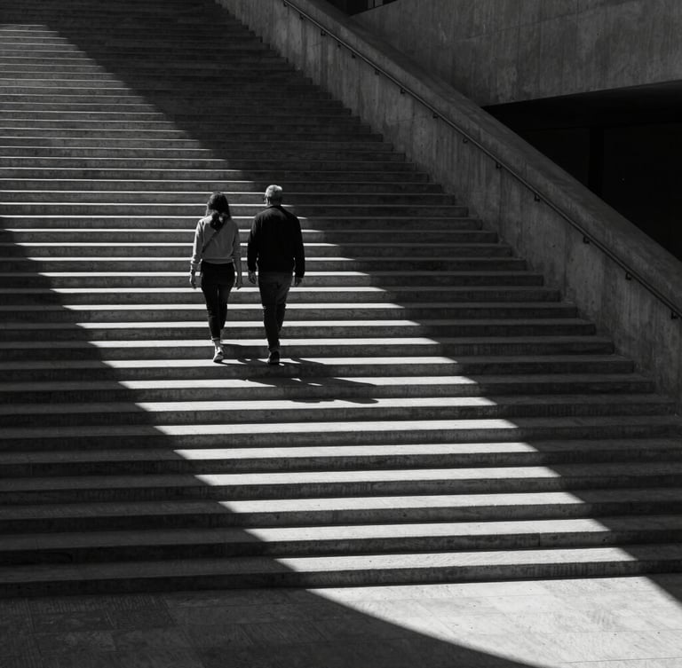 Wide cinematic environmental shot of a couple walking down a stark concrete staircase, dramatic high-contrast natural light casting long shadows, sharp monochrome, architectural framing.
