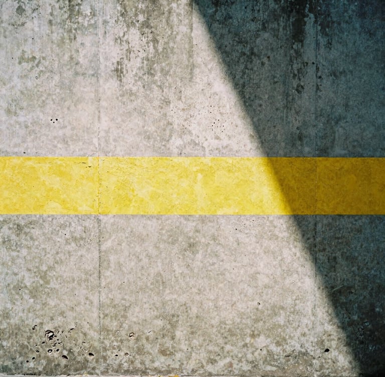Gritty editorial photography, high-contrast natural light, desaturated urban tones, sharp 35mm film look. Close-up of a weathered concrete wall in San Luis Potosí, marked with a bright industrial yellow stripe, sharp diagonal midday shadows.