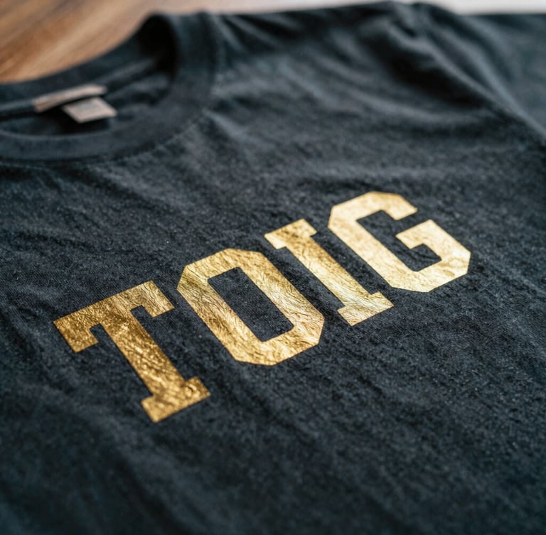 Macro close-up of custom metallic gold printing on a deep charcoal combed cotton t-shirt, showcasing the fine fabric texture and crisp print edges under warm window light, shallow depth of field, 35mm.