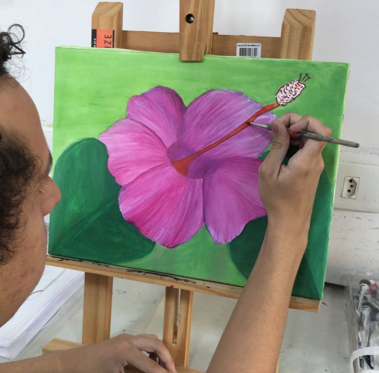 Artist painting a pink hibiscus flower on a green canvas using a fine brush and wooden easel.
