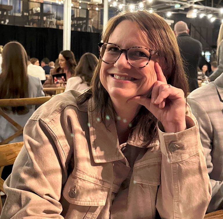 Smiling woman with glasses wearing a tan corduroy jacket at a dinner event with string lights.