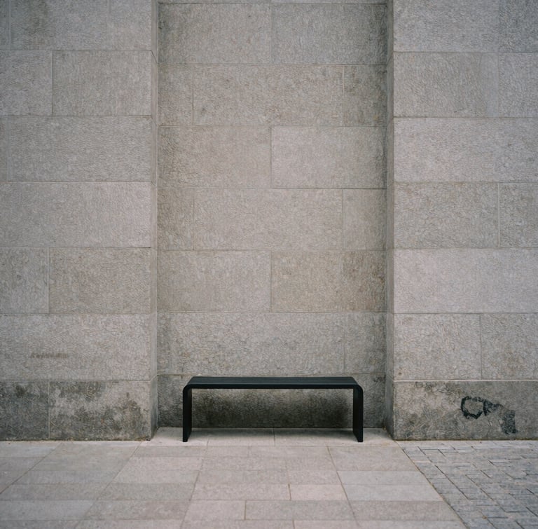 Architectural detail of a quiet Copenhagen courtyard, soft overcast light, textured gray stone walls, a single minimalist black metal bench, desaturated tones, 35mm film grain.