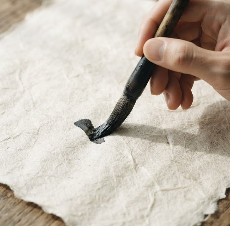 Extreme close-up flat lay of hands holding a wet bamboo calligraphy brush over raw, textured Xuan paper, soft natural side-light casting gentle shadows.