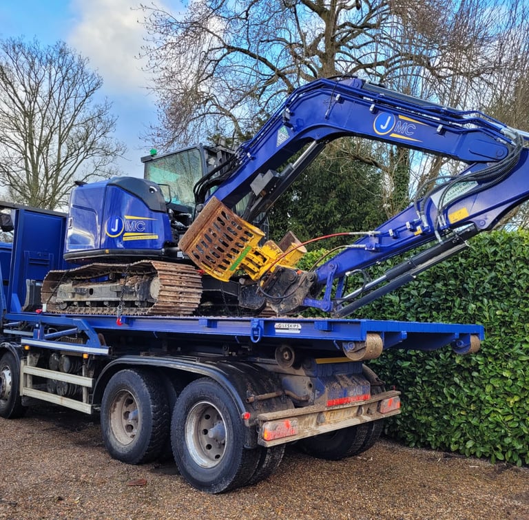 A blue JMC tracked excavator loaded onto a heavy-duty flatbed transport truck parked outdoors.