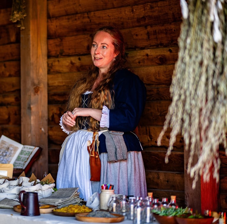a woman in 18th century clothing stands behind a table covered in herbal remedies