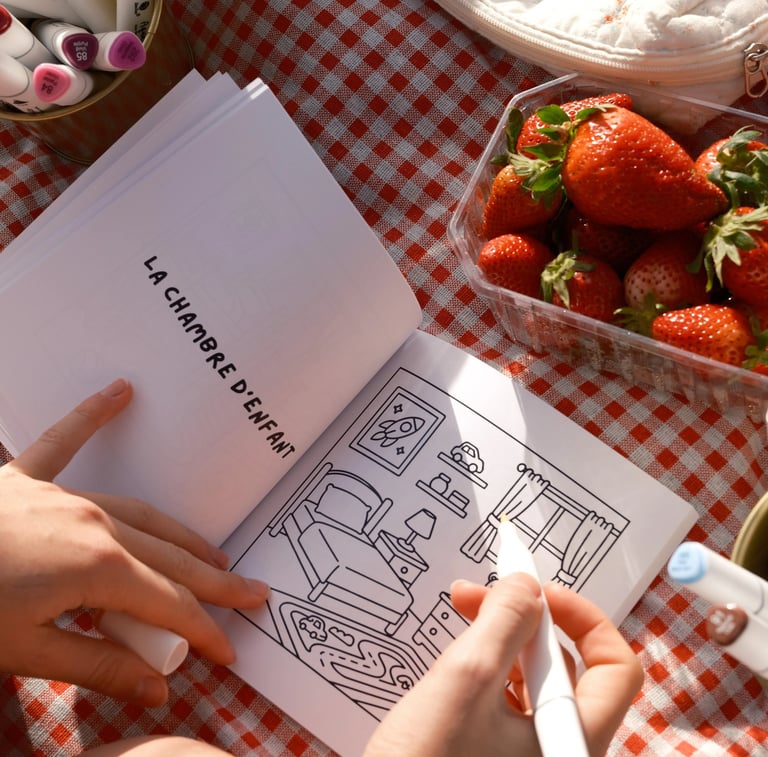 Une main coloriant une scène de chambre dans un livre de coloriage, à côté de fraises fraîches