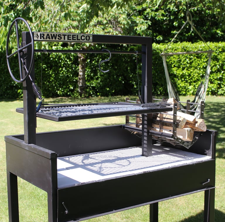 Custom black metal Argentine Santa Maria grill with flywheel and wood fire basket on a green lawn.