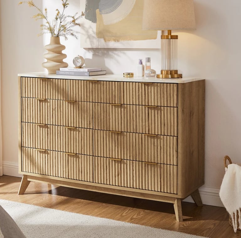 Modern mid-century 8-drawer fluted wood dresser with gold handles and a marble top in a bright bedroom.