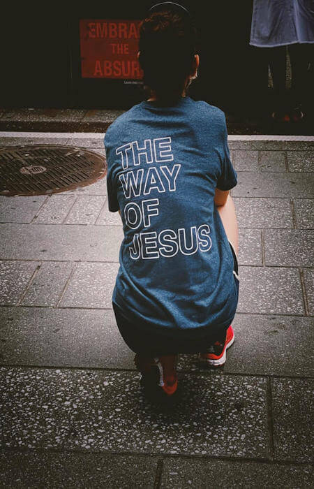 Person squatting on pavement wearing a shirt that reads “THE WAY OF JESUS” in bold white text.