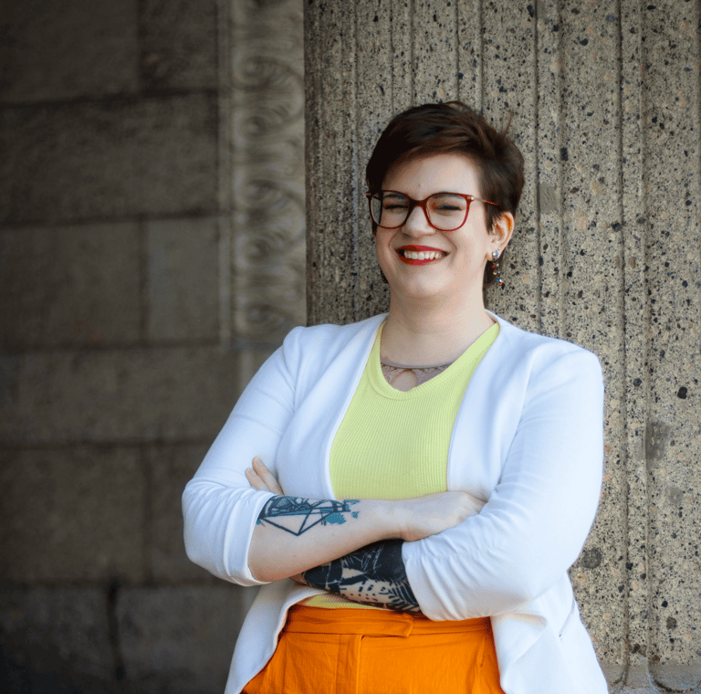ein Foto von Swana Schuchmann. Sie trägt einen weißen Blazer und lehnt sich an eine Säule, lachend.